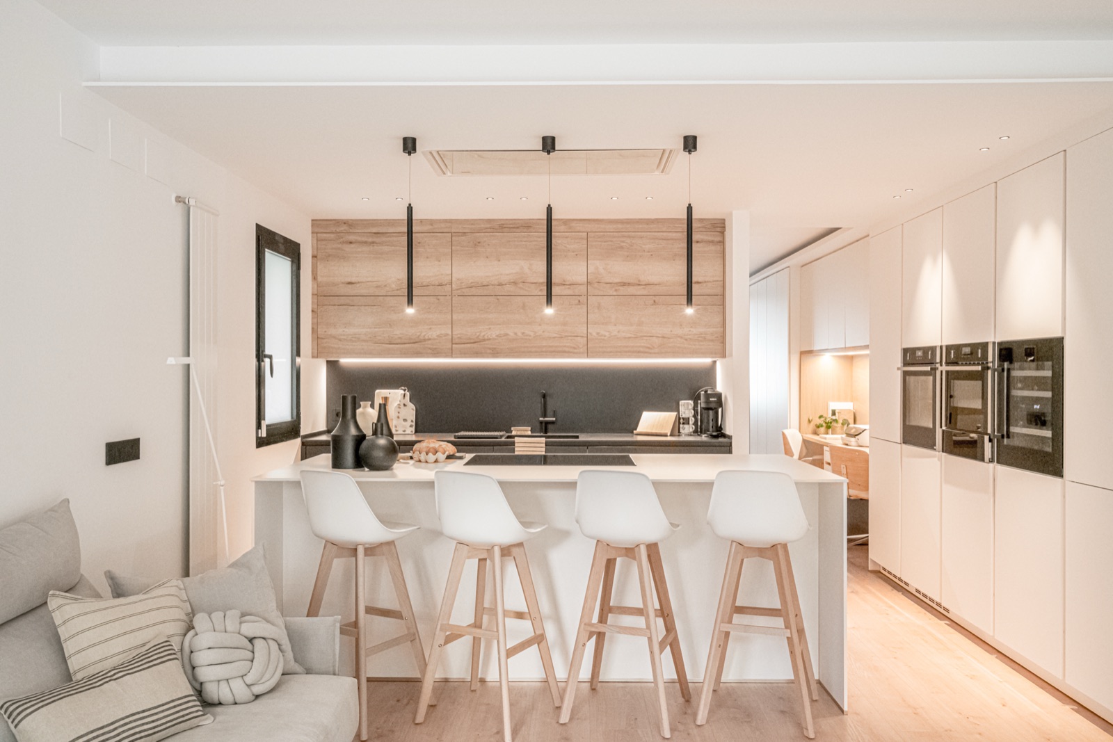 Cocina abierta con isla blanca, muebles de madera clara y lámparas colgantes negras