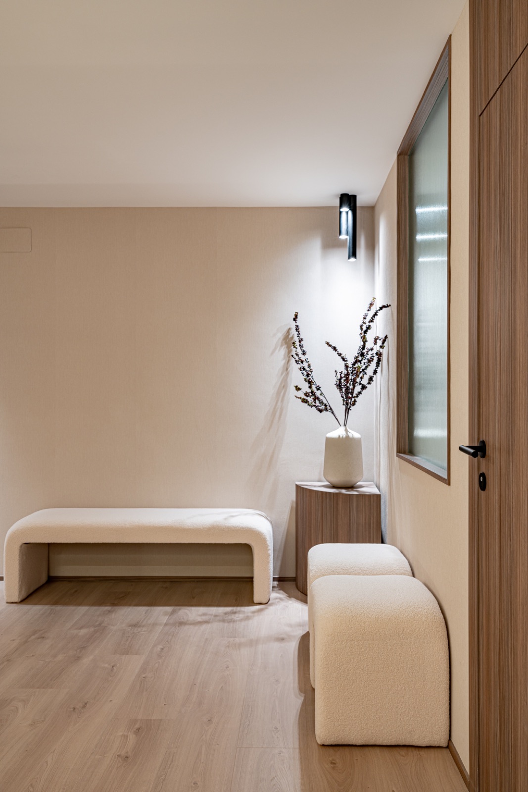 Sala de espera con bancos de bouclé blanco, jarrón con ramas secas y puerta de madera